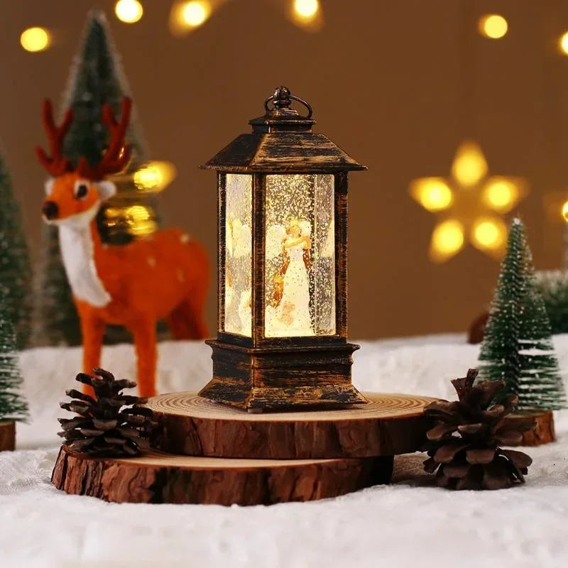 A rustic dark metal lantern with a warm glowing internal light sits on a round wood slice base, styled with pinecones and small artificial trees on a snowy white surface against a warm brown background with blurred yellow lights.
