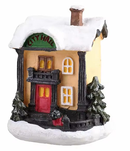 A small decorative village house figurine with a snowy roof and base, featuring a beige facade, black trim, a red front door, yellow glowing windows, and green trees on a white background.