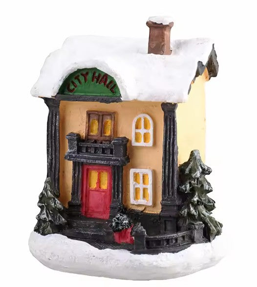 A small decorative holiday house figurine labeled 'CITY HALL' with a snowy roof, red front door, black columns, and dark green evergreen trees on a white snowy base.